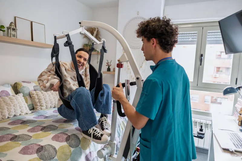 A caregiver helps a young man transfer from his bed using a patient lift at home