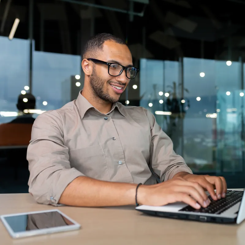 Portrait of Samir Rao, a smiling man with glasses and a short beard, working on a laptop in a glass-walled office