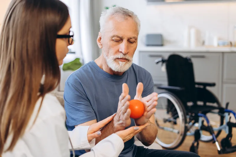 An older man squeezes a therapy ball during hand therapy, with a wheelchair behind him