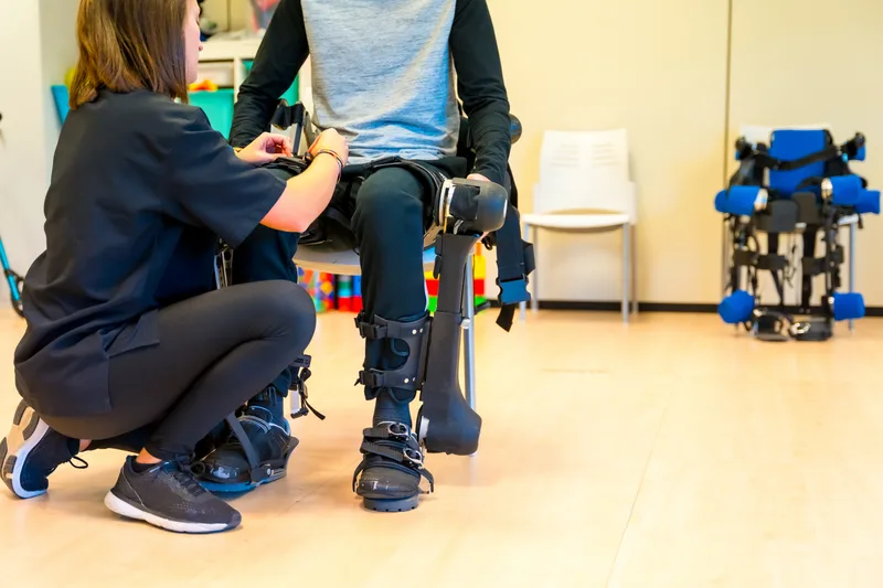 A physical therapist kneels to adjust the leg braces of a patient standing in a rehabilitation exoskeleton
