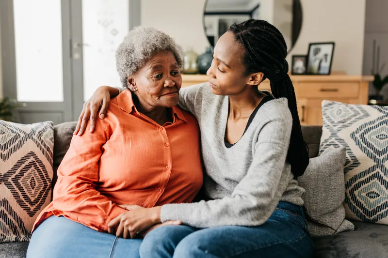 A young woman sits on a sofa with her arm around her mother, comforting her