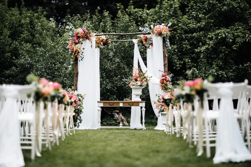 Garden ceremony set with white chairs, a draped floral arch, and a grass aisle between hedges