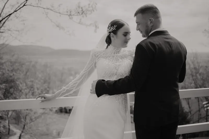 Bride in a beaded high-neck gown and groom in a dark suit standing at a white balcony rail, photographed in black and white with hills behind them