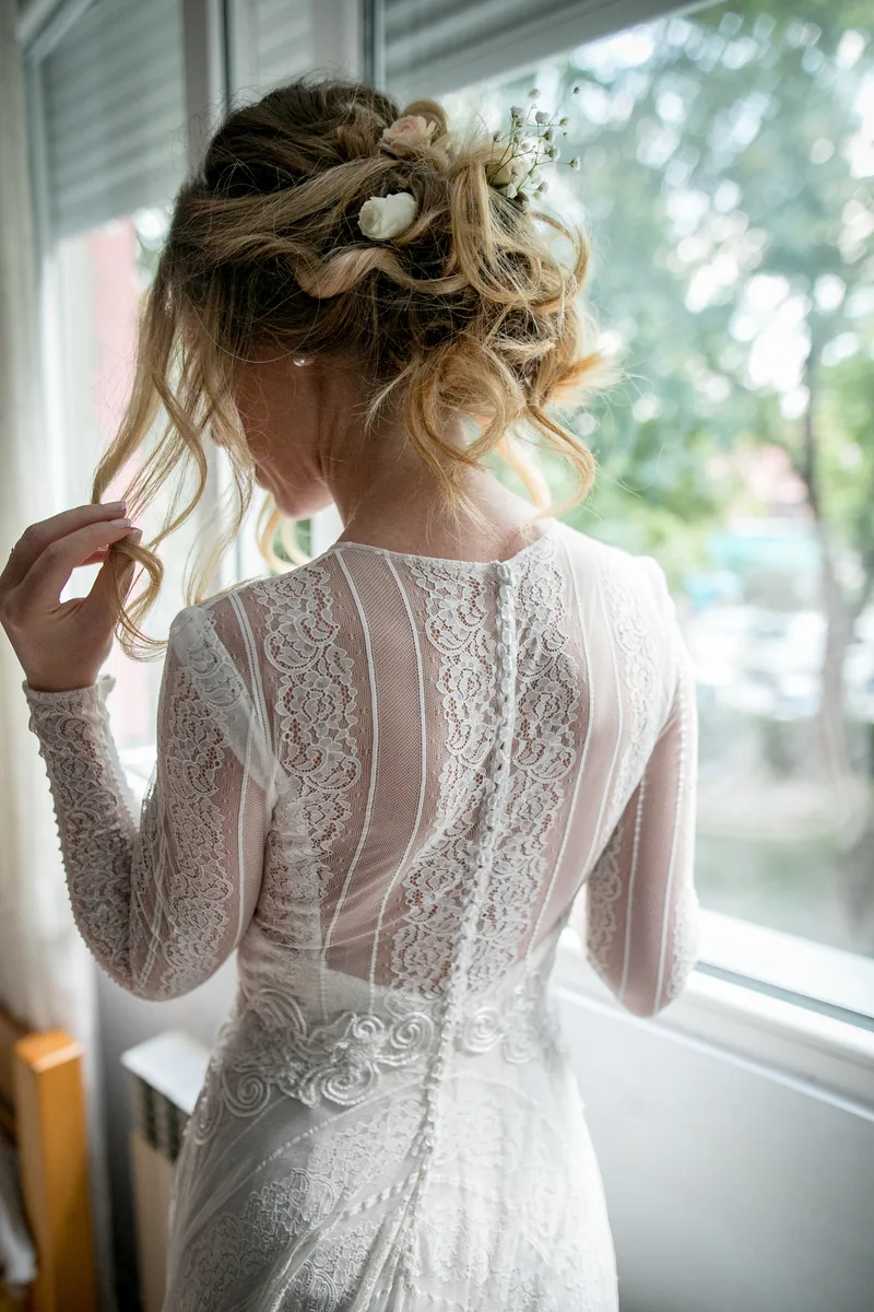 Bride photographed from behind in window light, showing the lace back of her gown and white flowers pinned into her updo