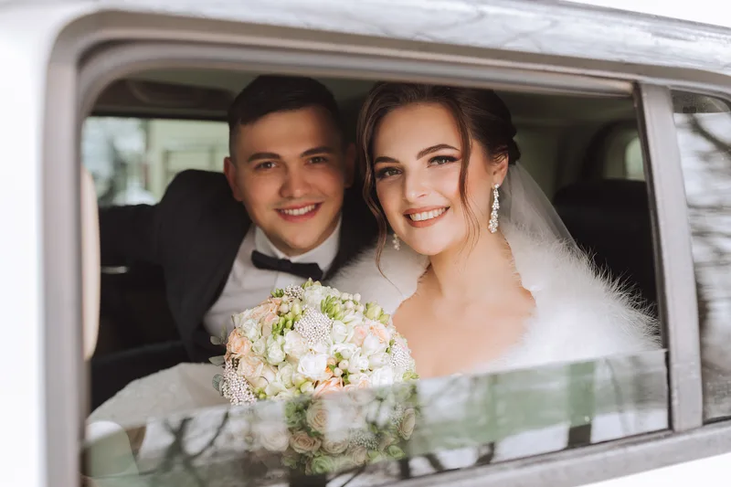 Smiling bride and groom looking out of a car window together, her bouquet of white and blush roses resting on the door