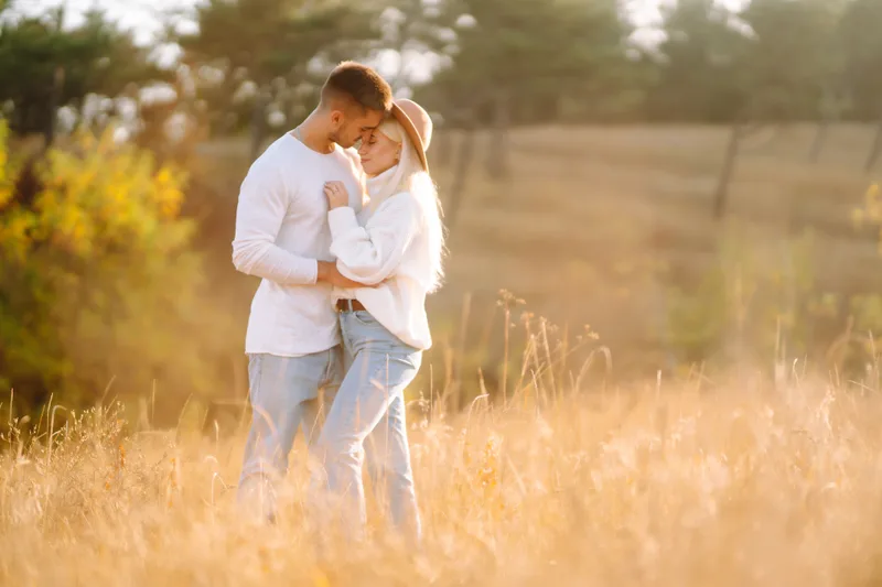 Couple in white sweaters embracing forehead-to-forehead in a golden backlit field, the woman wearing a brimmed hat
