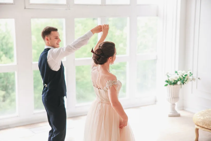 Groom twirling the bride in a light-filled room with tall white windows, her blush gown mid-turn