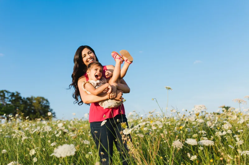 Mother lifting her laughing young daughter in a field of white wildflowers under a clear blue sky