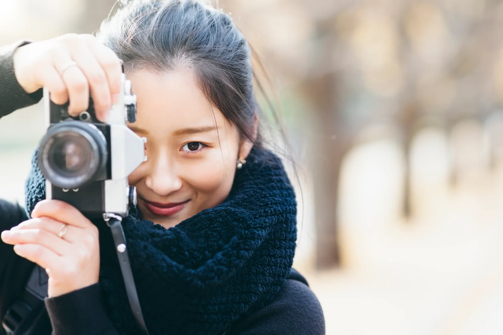 Emi Sato holding a film camera up to her eye, one dark eye visible past the lens, wrapped in a navy knit scarf