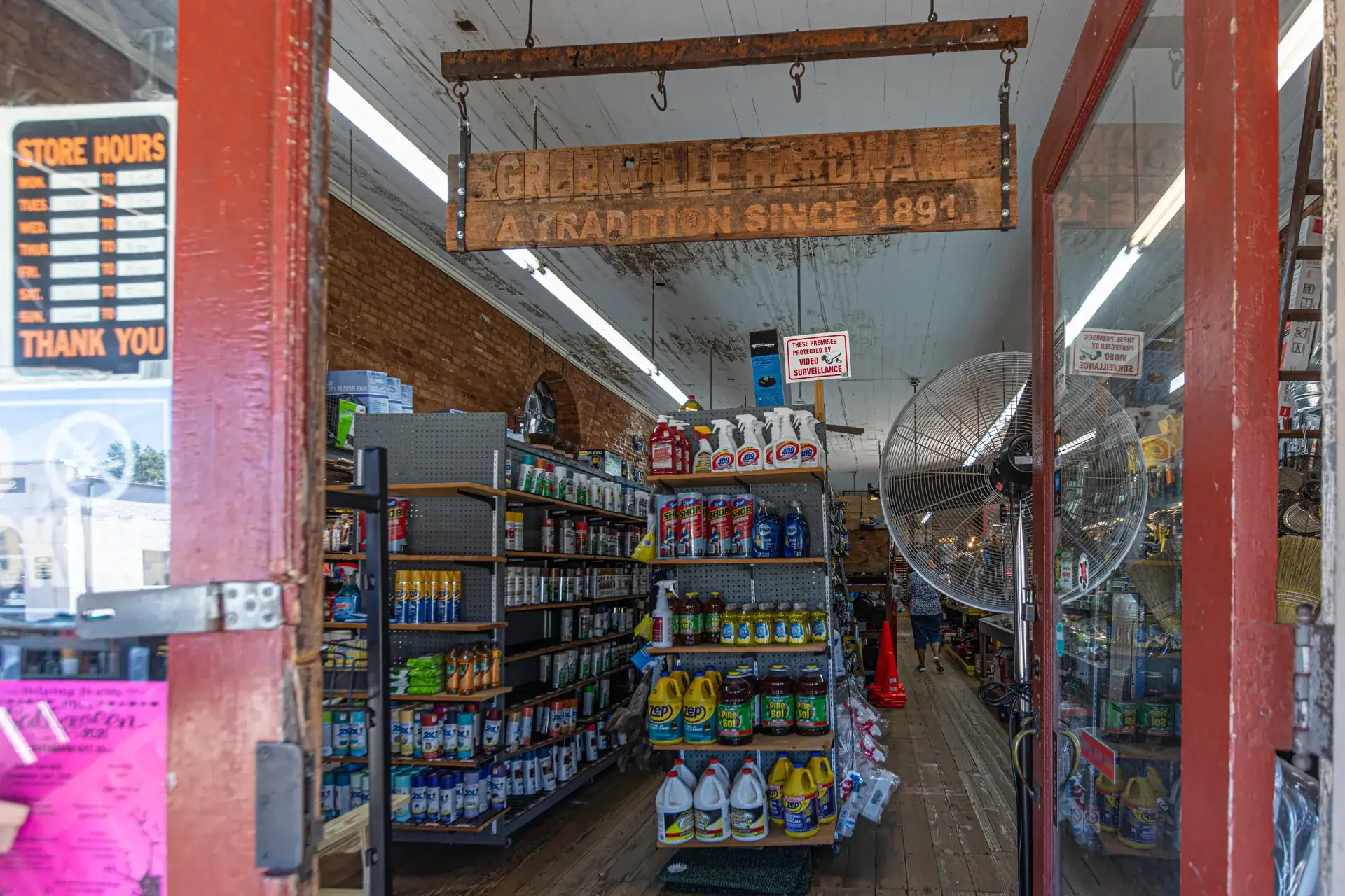 The open front door of a long-standing main-street hardware store — red-painted frame, a weathered wooden sign hanging from the ceiling, and stocked aisles running deep under old brick walls
