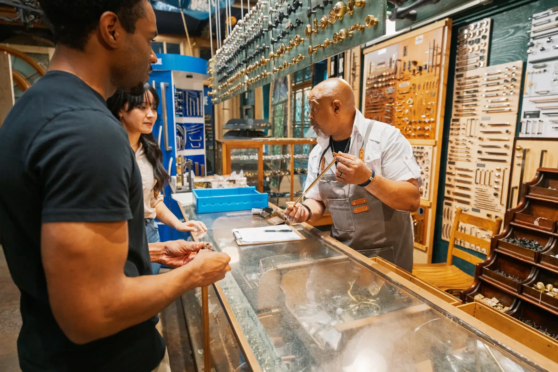 A counterman in a work apron measuring a length of brass hardware for two customers at a glass parts counter, walls of drawer pulls and hinges behind him