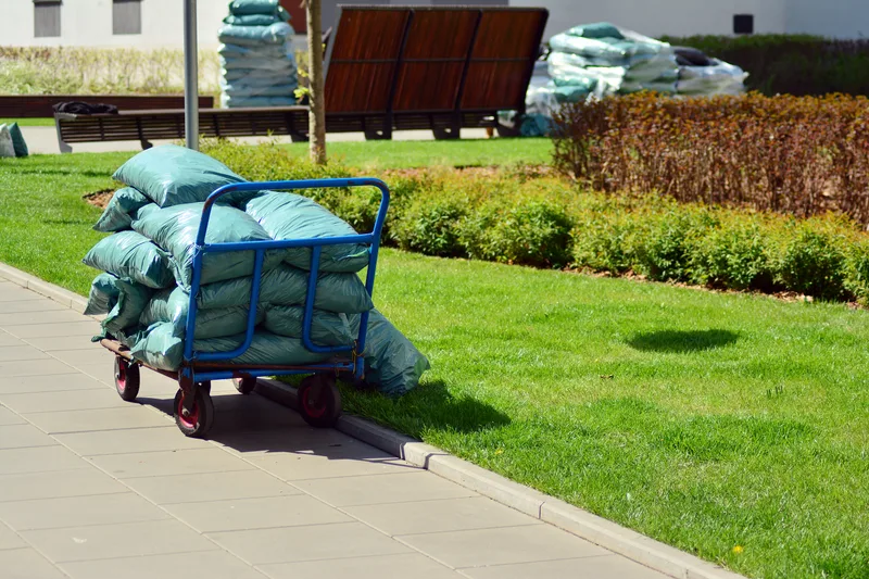A blue garden cart stacked with bagged mulch beside a lawn and hedge