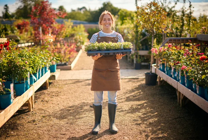 A Loamwood staff member in a canvas apron carrying a tray of seedlings down a garden centre aisle lined with flowering plants
