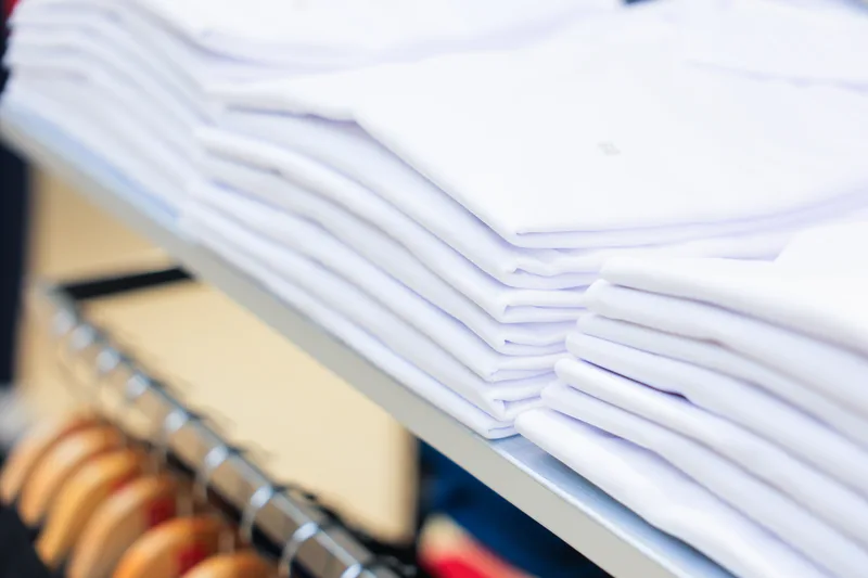 Stacks of laundered white shirts, folded and tissue-wrapped, on a shelf above a row of wooden hangers