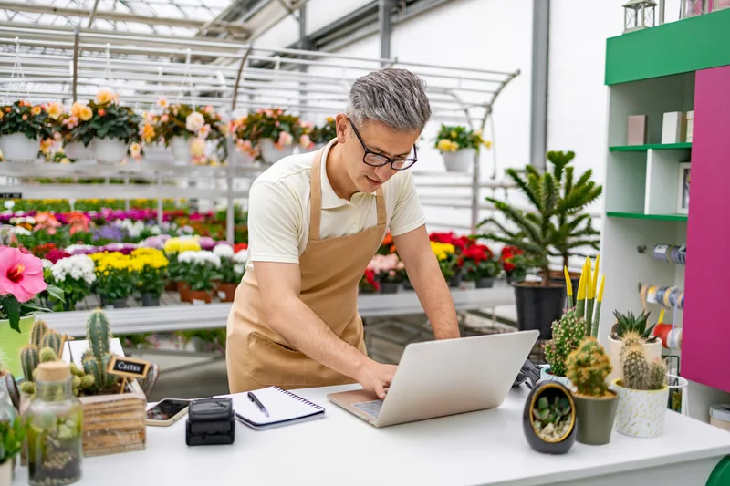 A Tansy florist checking the day's orders on a laptop among buckets of potted flowers and cacti