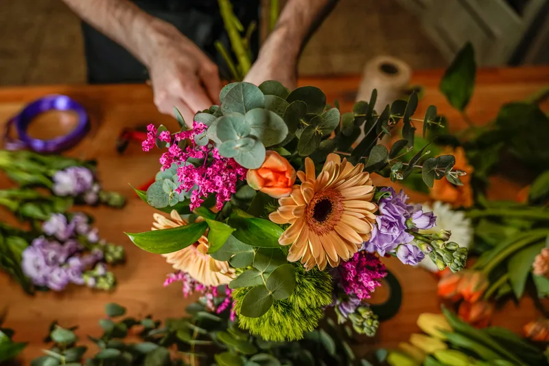 Overhead view of a Tansy designer arranging a bright bouquet of gerberas, stock and eucalyptus on the wooden bench