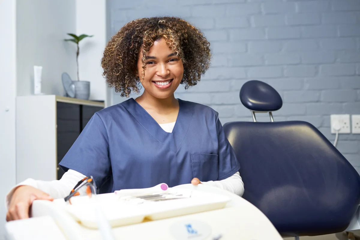 A smiling hygienist seated beside a navy dental chair with her instrument tray prepared