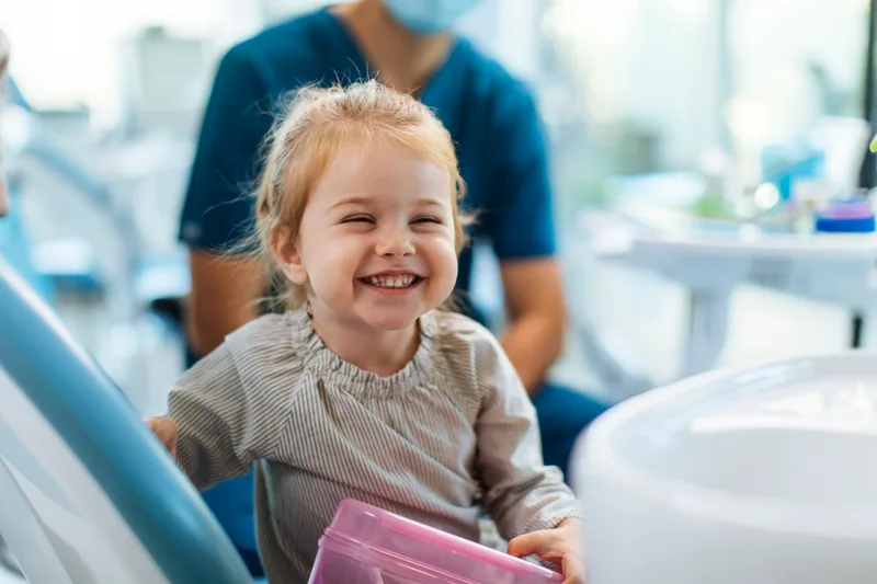 A toddler mid-laugh in the dental chair while a masked clinician looks on behind her