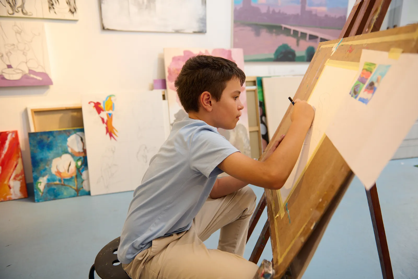 A young student painting at an easel while an art teacher looks on in a colorful, supply-filled kids art studio