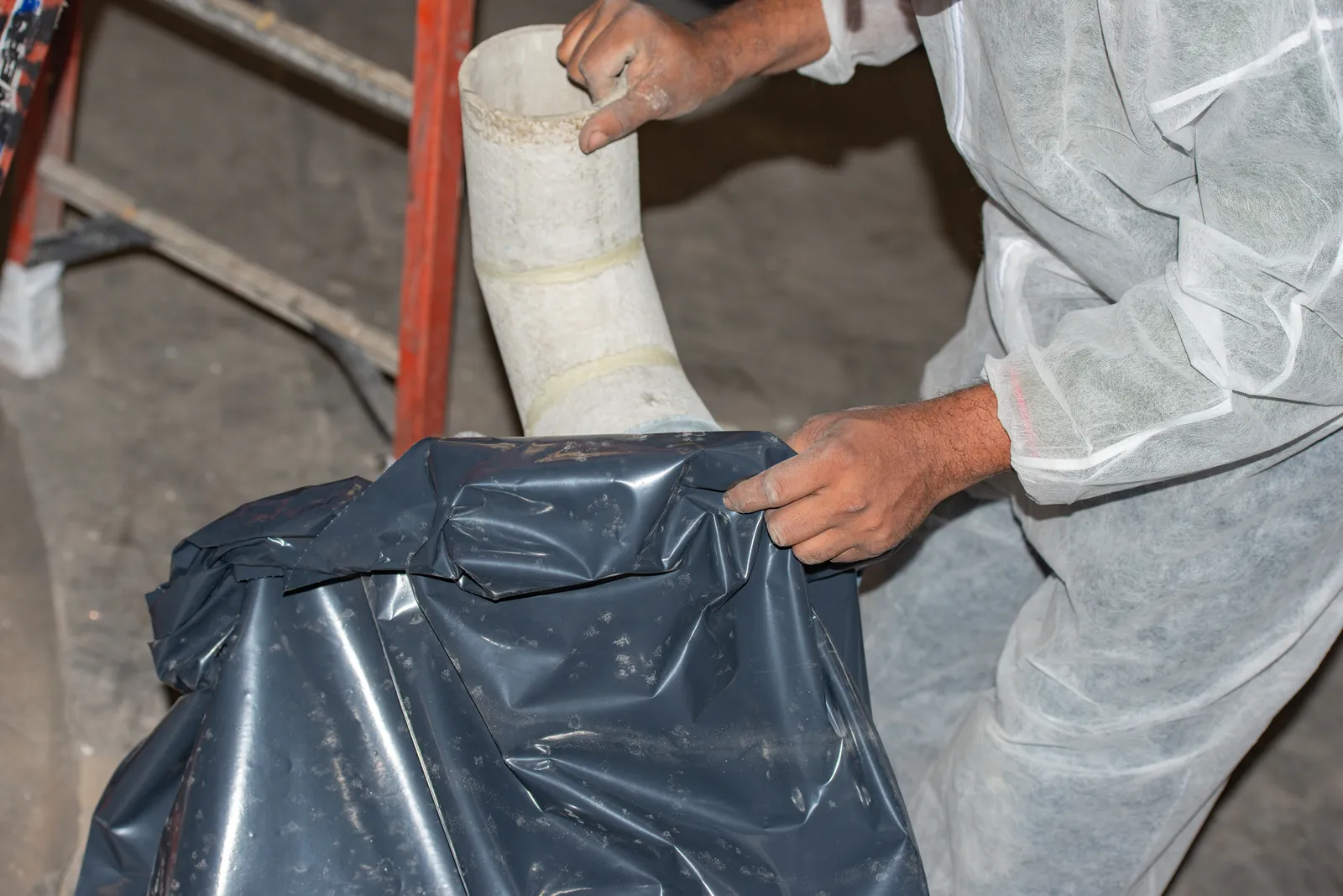 Certified technician in a full-body protective suit and respirator removing asbestos insulation from an overhead pipe in a work area sealed with plastic sheeting