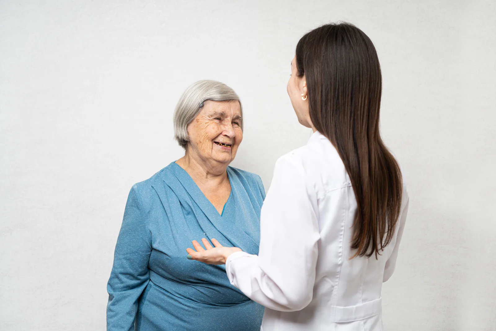 A caregiver holds hands with a smiling senior resident in a sunlit common room at an assisted-living community