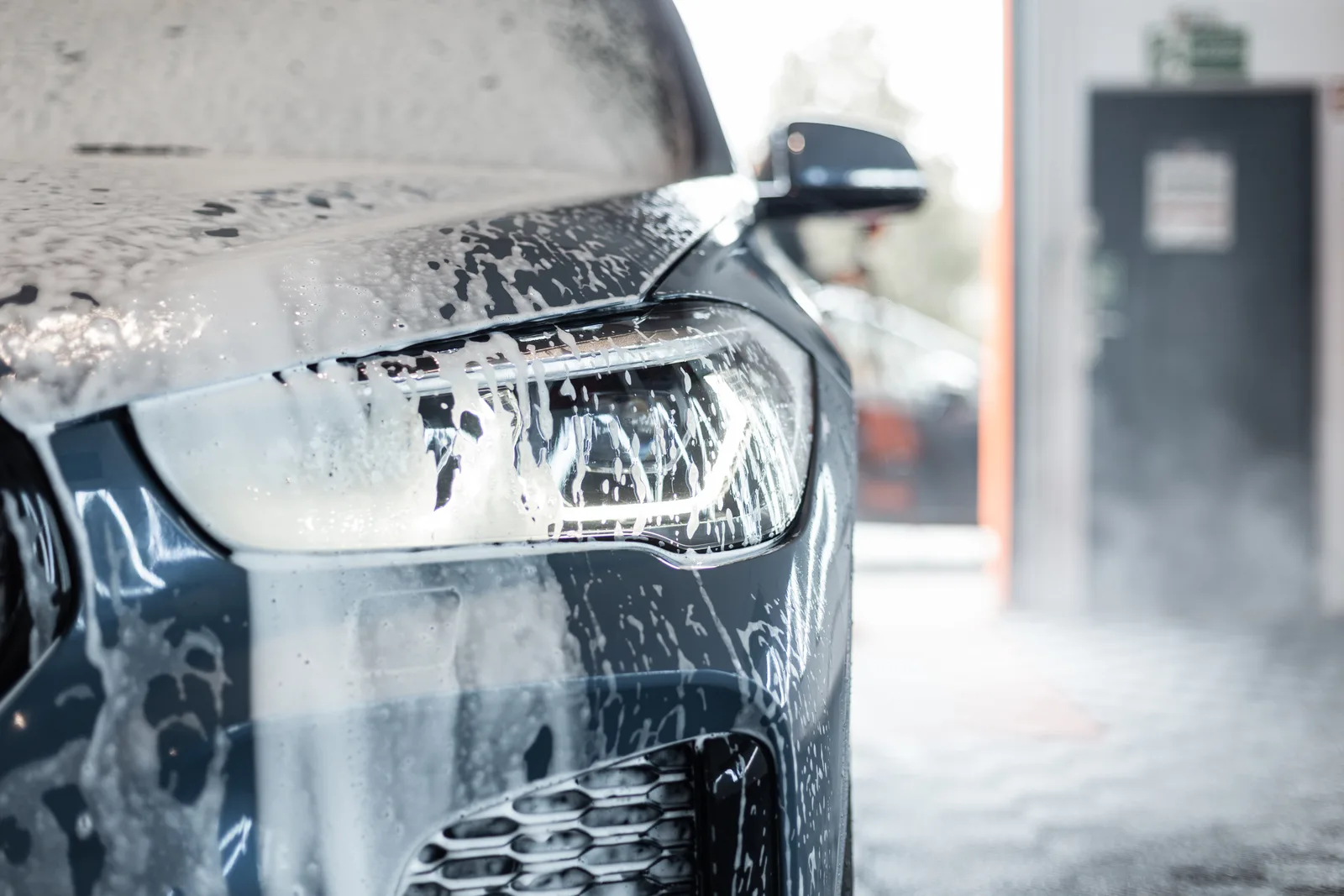 Sedan rolling through a tunnel car wash as foam and soft cloth brushes cover its roof and doors