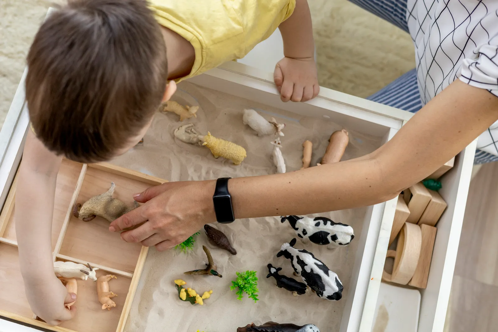 A young child arranging toy figures in a sand tray while a play therapist sits nearby during a session