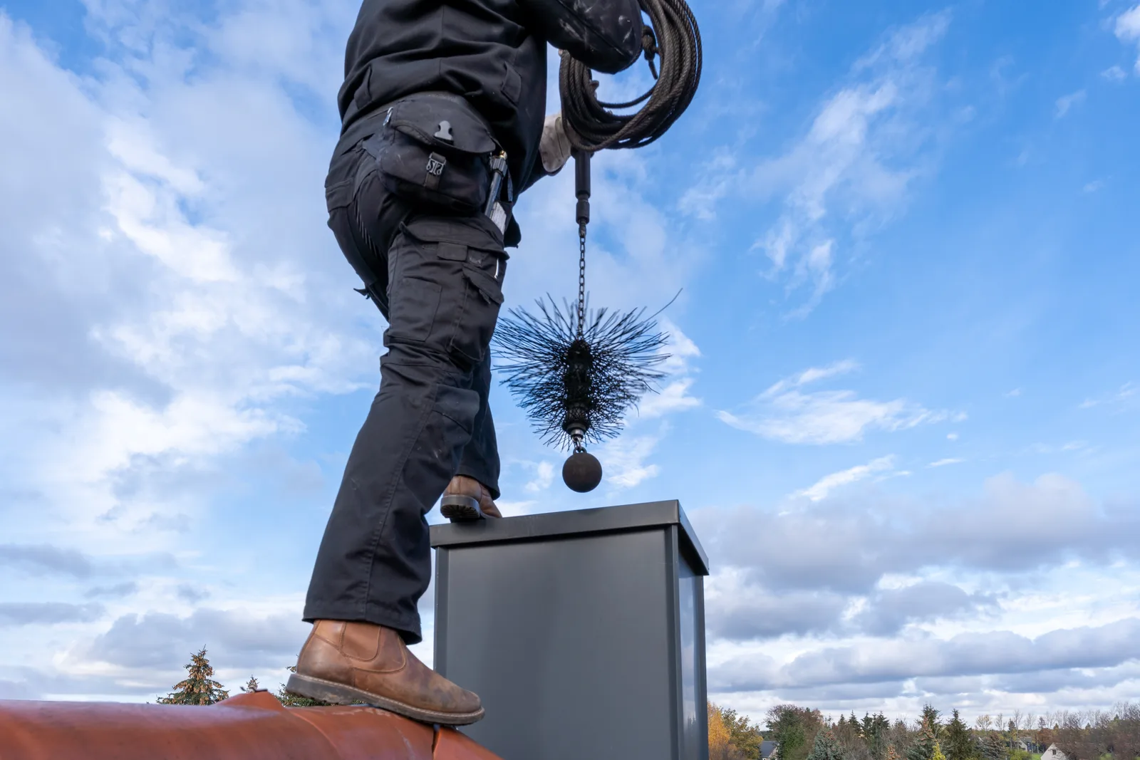 Chimney technician sweeping a brick chimney with a rod and brush from the rooftop of a family home