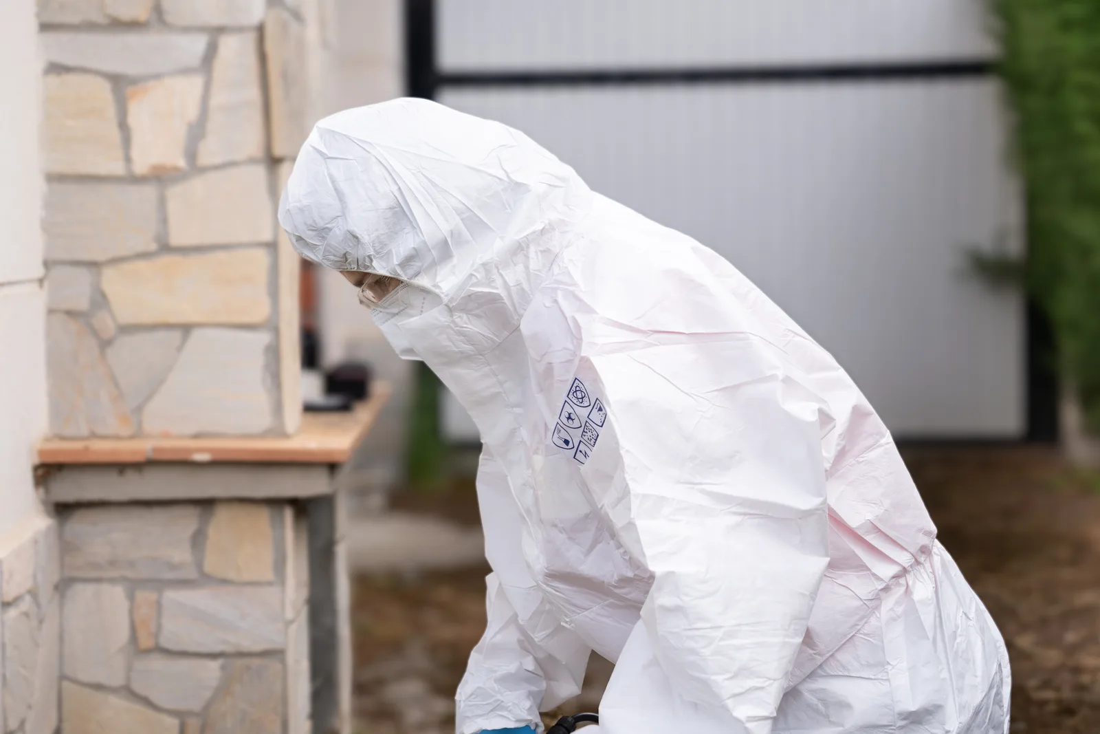 A biohazard remediation technician in a full protective suit and respirator carrying cleaning equipment into a home for a decontamination job