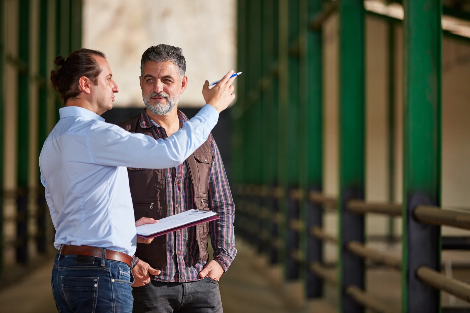 Equine attorney reviewing a horse purchase agreement and pre-purchase veterinary exam report with a client in a stable aisle, horses visible in the stalls behind them