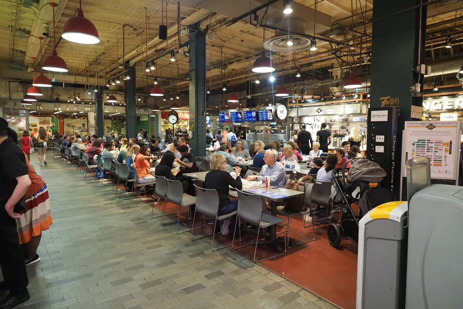 Guests seated at long communal tables inside a busy food hall, with lit vendor stalls and menu boards lining the concourse behind them