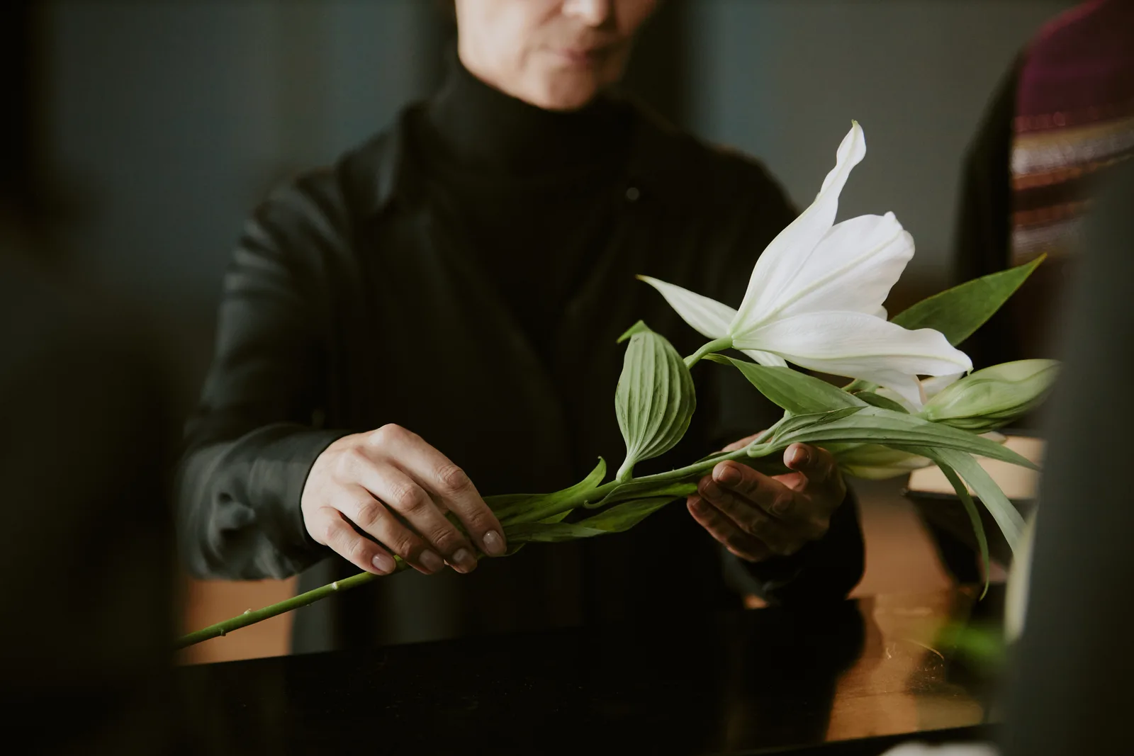A funeral director in a dark suit arranges white lilies on a memorial table inside a warmly lit funeral home chapel