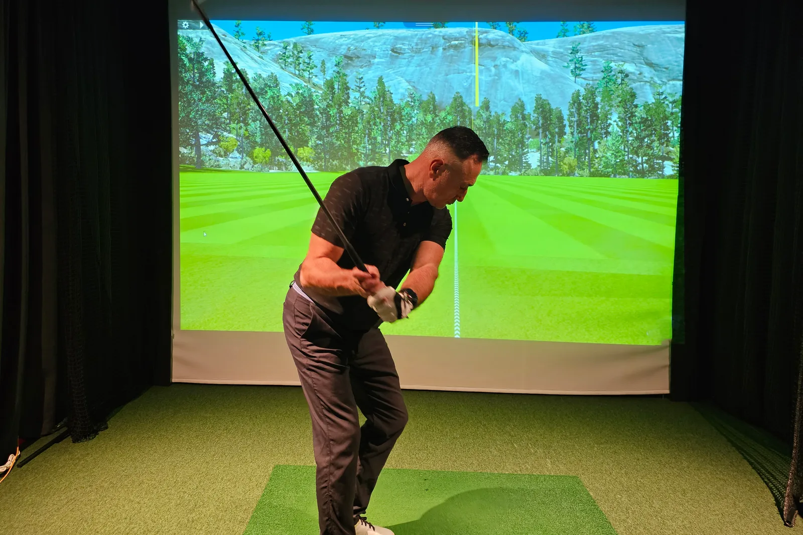Golfer swinging a driver inside an indoor golf simulator bay, with the shot tracked on a large projector screen