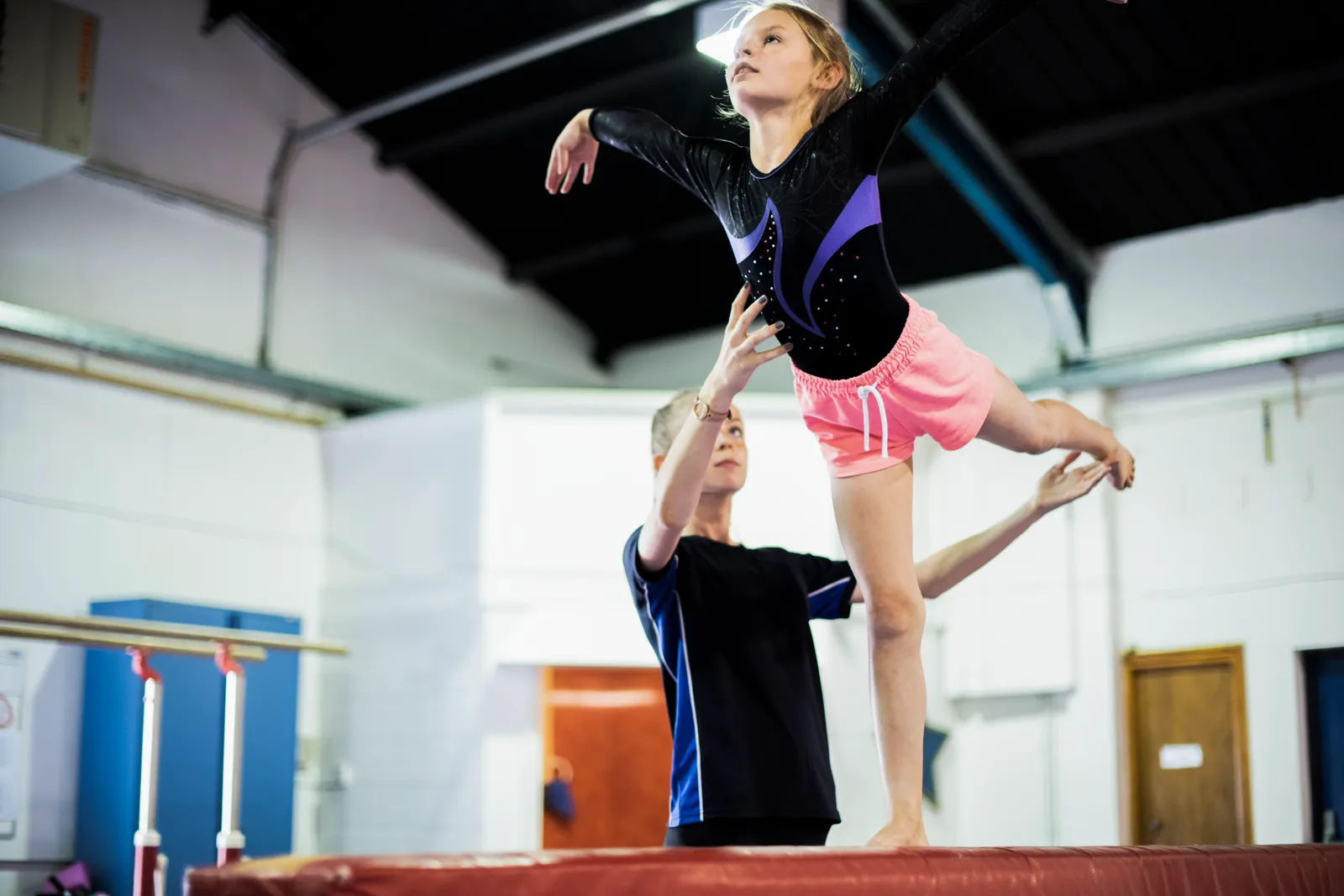 Young gymnast balancing on the beam while a coach spots her in the gym