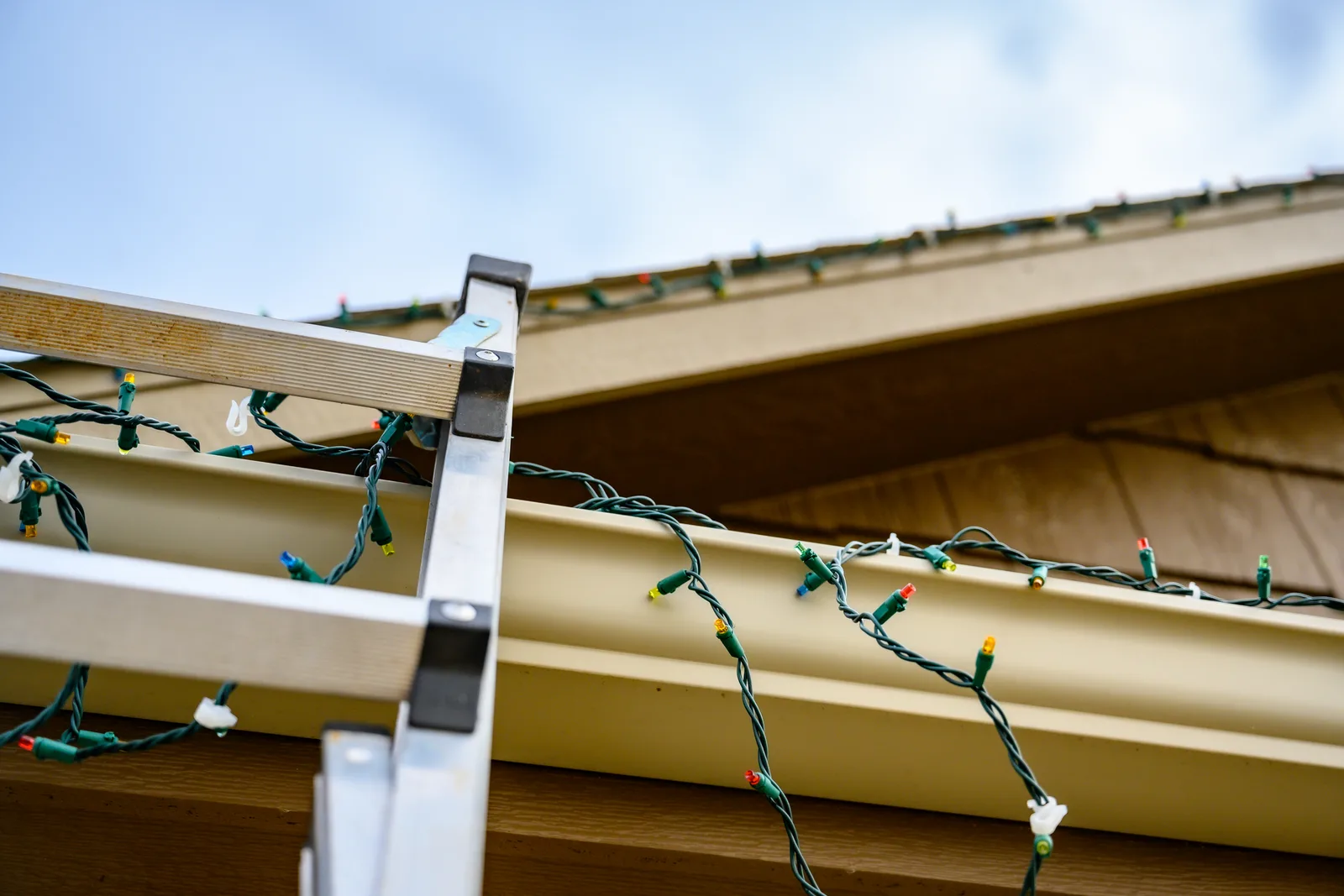 Installer on a ladder hanging warm-white string lights along a home's roofline at dusk