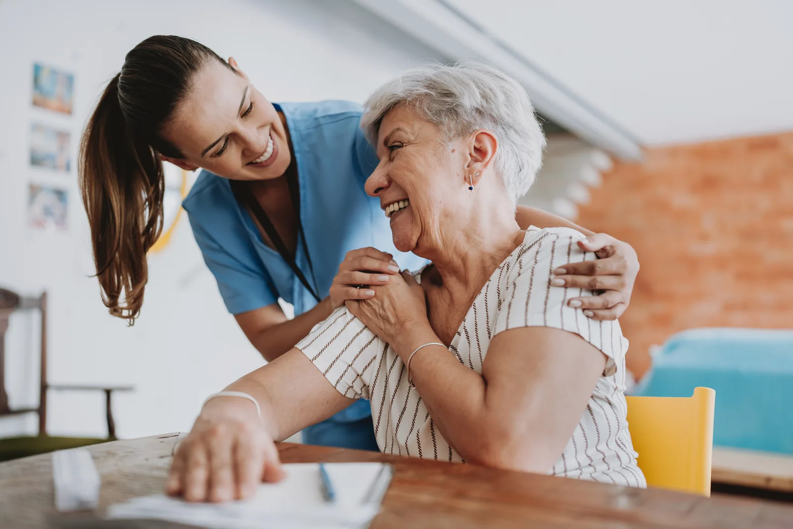 A home caregiver shares a warm laugh with a smiling senior woman on the couch in her own living room