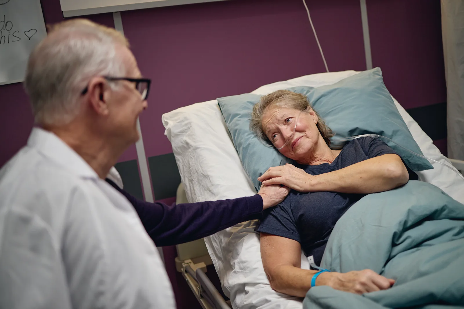 A hospice nurse gently holds the hand of an elderly patient at the bedside while a family member looks on