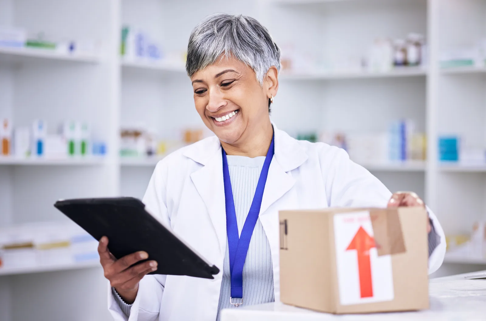 An independent pharmacist handing a prescription bag to a patient across the counter of a neighborhood pharmacy