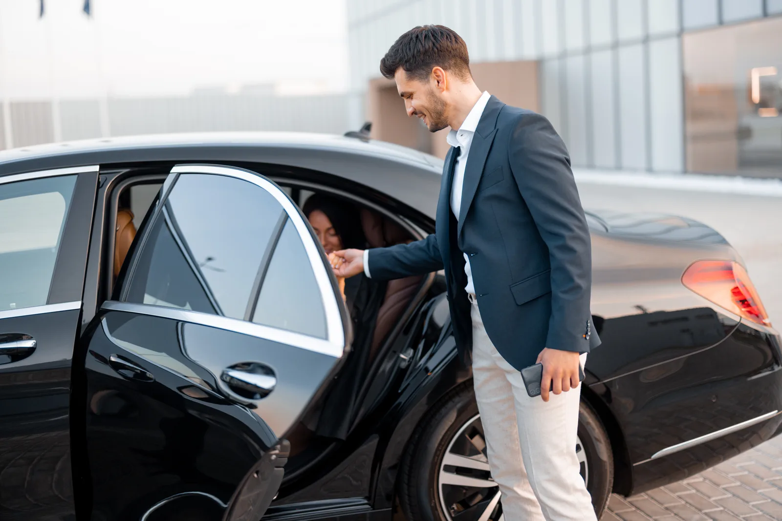 Uniformed chauffeur opening the rear door of a black limousine for a client at the curb
