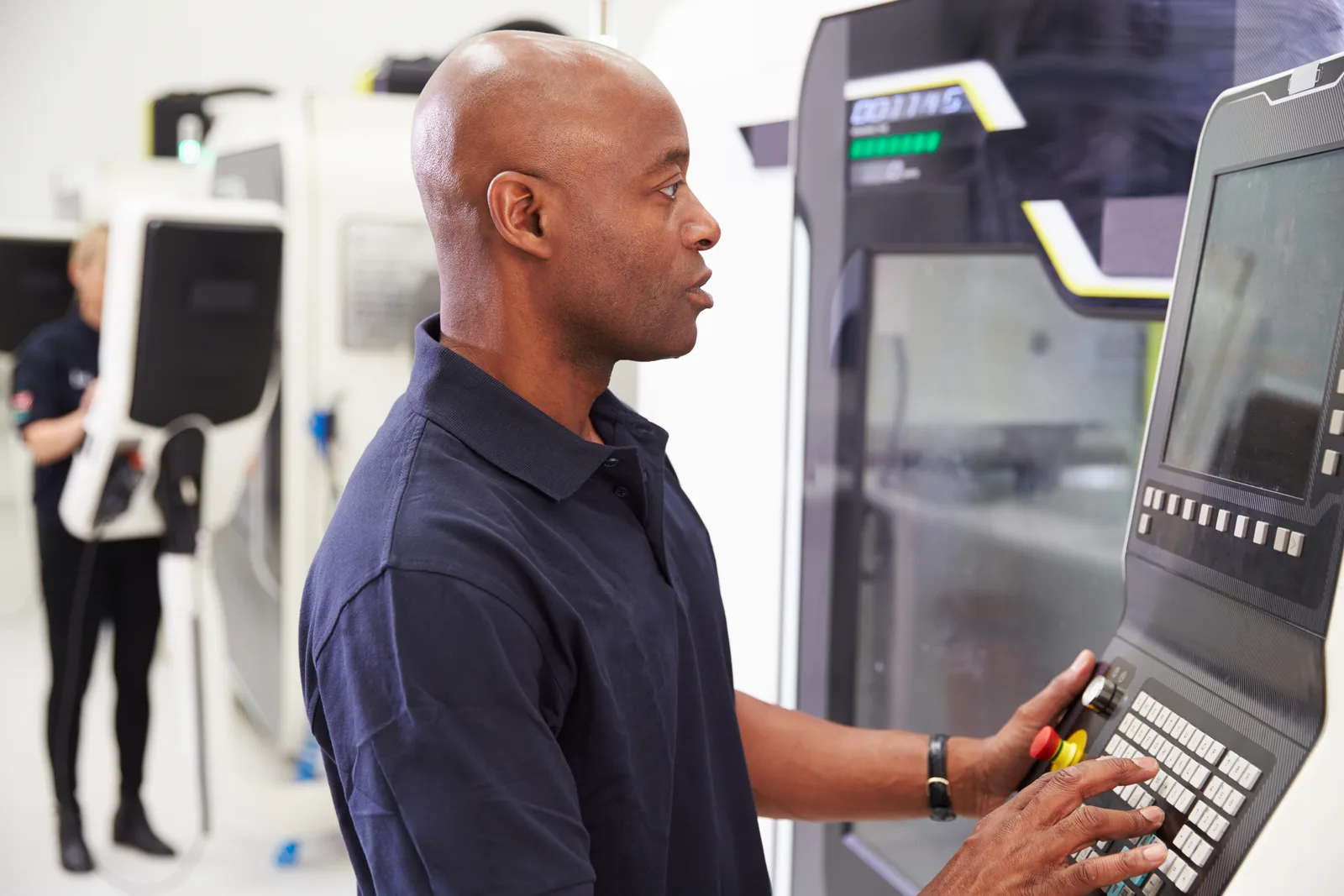 Machinist inspecting a precision metal part next to an active CNC milling machine on the shop floor