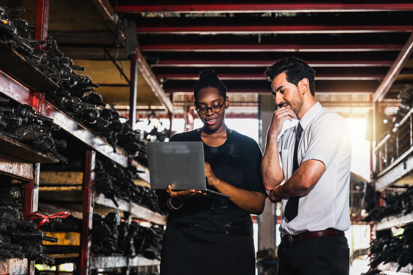 Manufacturing team reviewing data on a laptop together in a warehouse aisle lined with parts storage racks