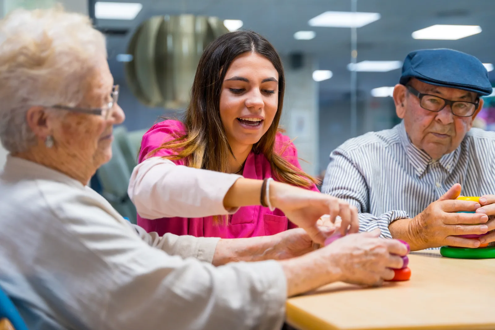 A caregiver sits close beside a smiling senior resident, guiding a hands-on memory activity in a secured memory care community common room