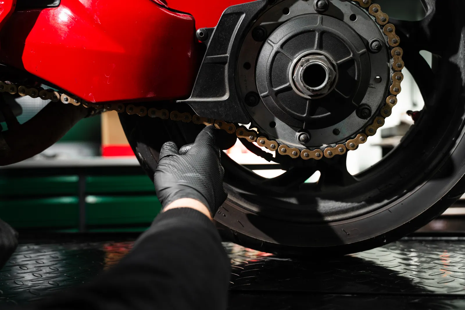 Mechanic's gloved hands adjusting a motorcycle's chain and rear sprocket on a lift in a repair shop bay
