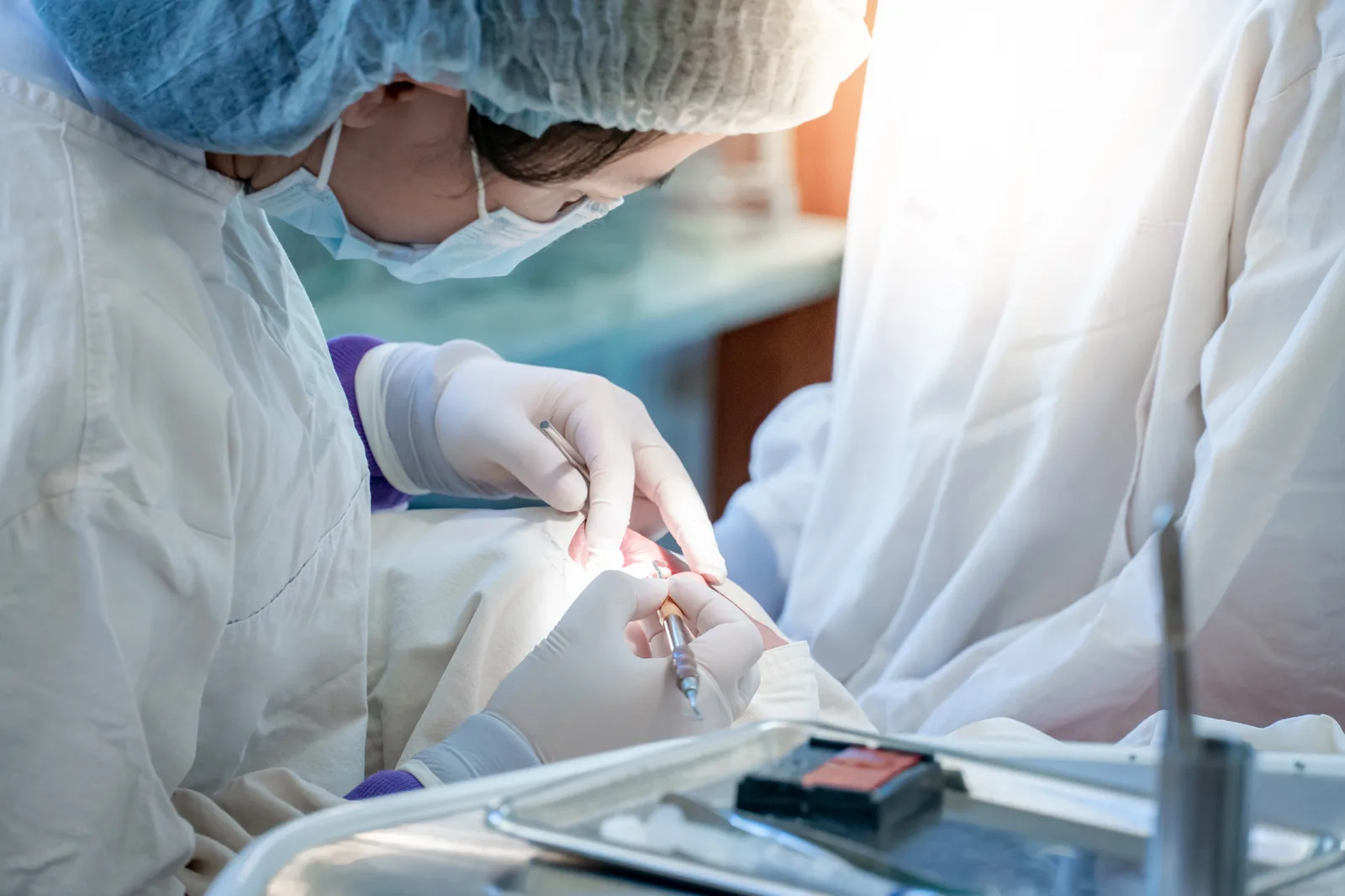 Masked oral surgeon in cap and gloves operating inside a patient's mouth, instrument tray in the foreground
