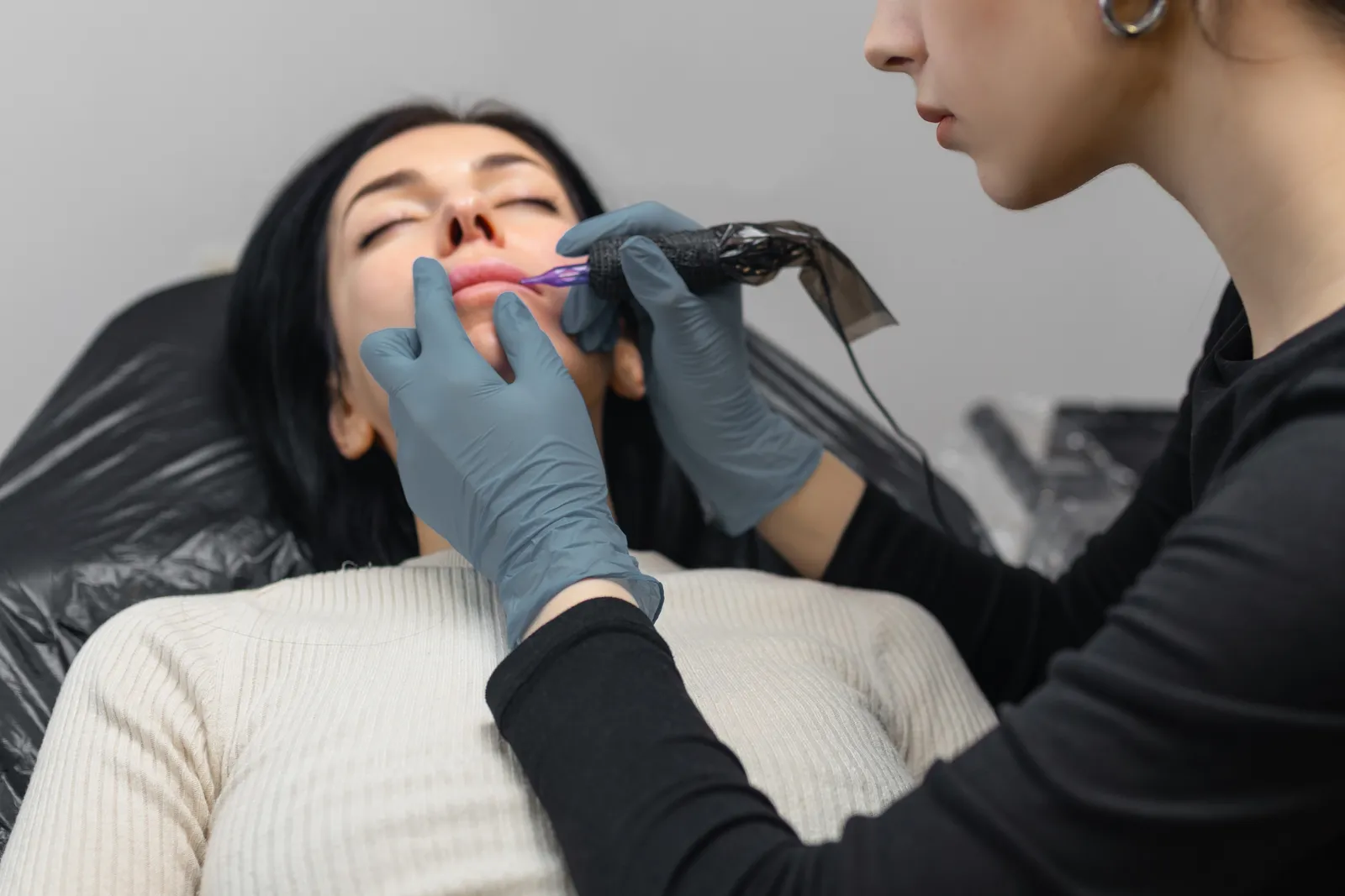 Permanent makeup artist applying lip blush pigment to a client's lips at a treatment table in a PMU studio