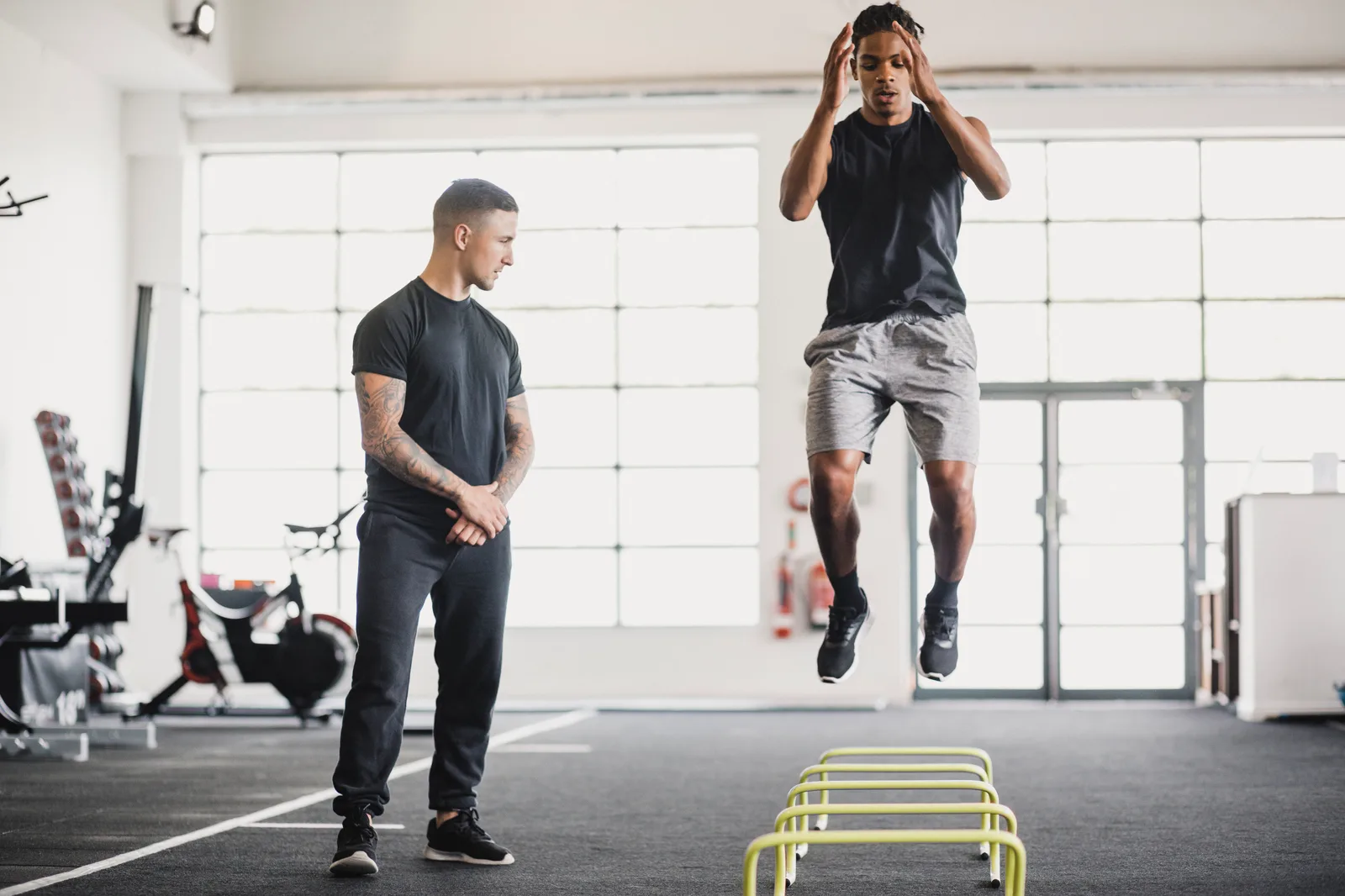 Personal trainer watching his client leap over agility hurdles during a one-on-one session in a bright gym