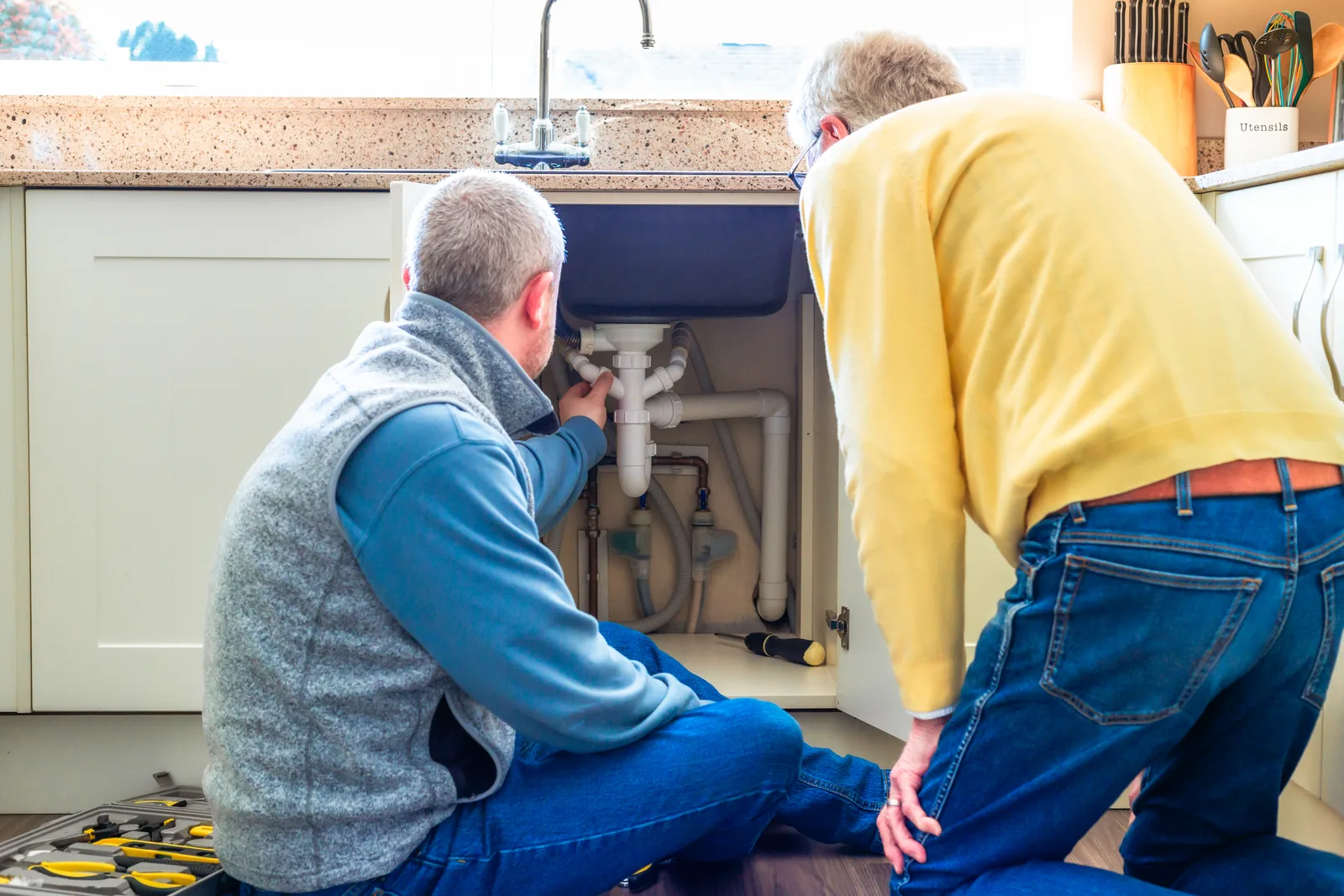 Plumber fitting a new drain trap under a kitchen sink while the homeowner looks on