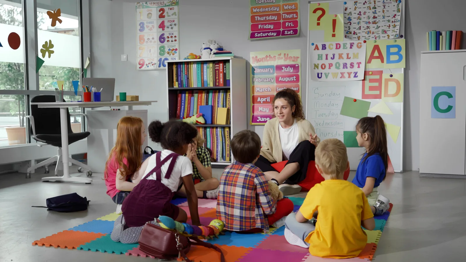 A preschool teacher reads a picture book to toddlers gathered on the classroom rug