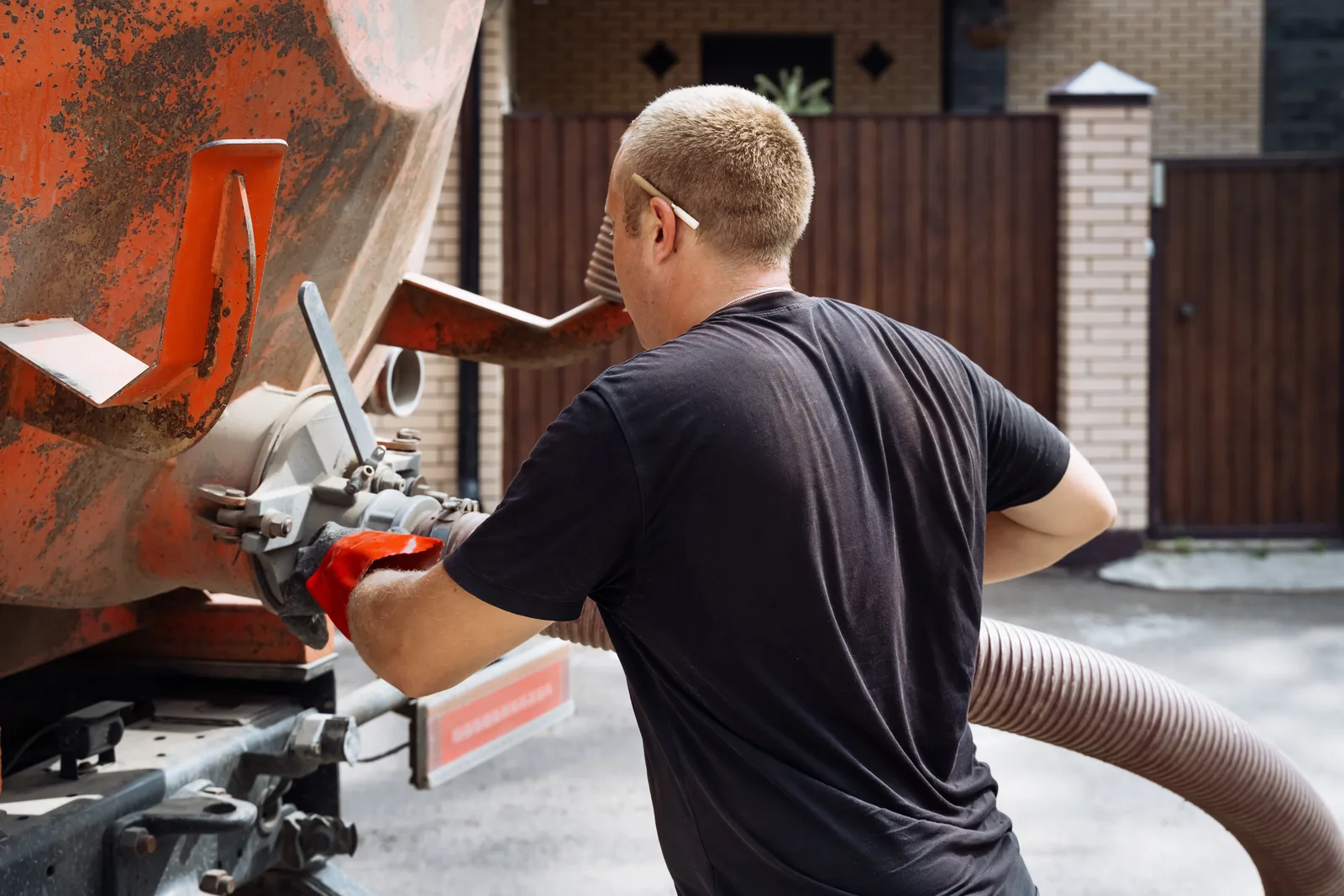 Septic technician guiding a vacuum-truck hose to pump a residential septic tank in a rural backyard