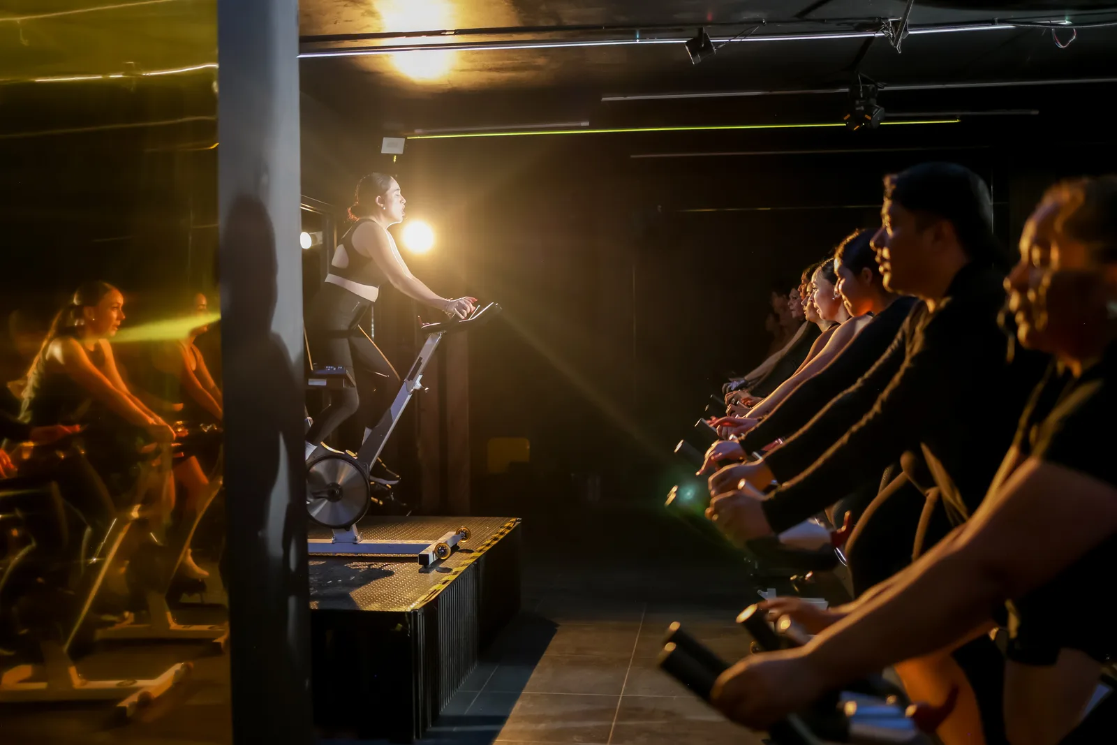 Riders on stationary bikes in a dimly lit cycling studio, following an instructor mid-class